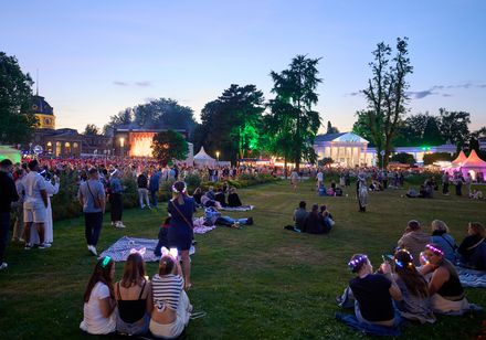 Die Parklichter zur blauen Stunde. Tausende Besucher genießen die Parkatmosphäre im Rahmen der Parklichter am Samstag zum Veranstaltungsmotto "Feel the 90s". Zahlreiche Menschengruppen liegen mit Picknickdecken auf dem Rasen und lauschen dem Konzert der Söhne Mannheims im Hintergrund.