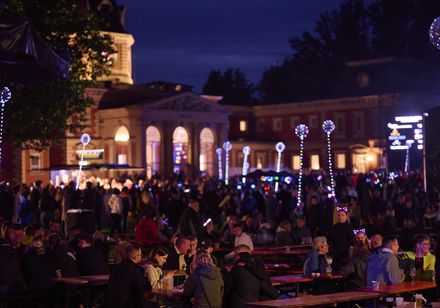 Menschenmengen an Sitzgarnituren bei den Parklichtern 2025 zur Griechischen Nacht, beleuchtete Luftballons und Badehaus II im Hintergrund bei Nacht.